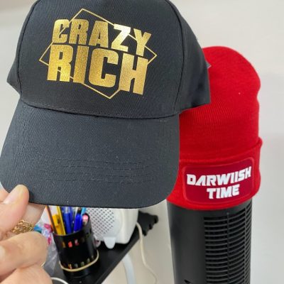 Black baseball cap with bold yellow text “CRAZY RICH,” held in front of other caps including a red one labeled “Darwin Time.”